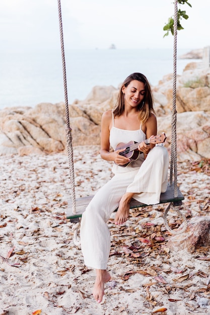 Romantica giovane donna caucasica calma felice con ukulele sulla spiaggia rocciosa tropicale al tramonto
