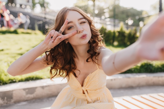 Romantica giovane donna dai capelli rossi che fa selfie nel fine settimana estivo. Foto all'aperto della ragazza positiva allo zenzero che cattura maschera di se stessa.