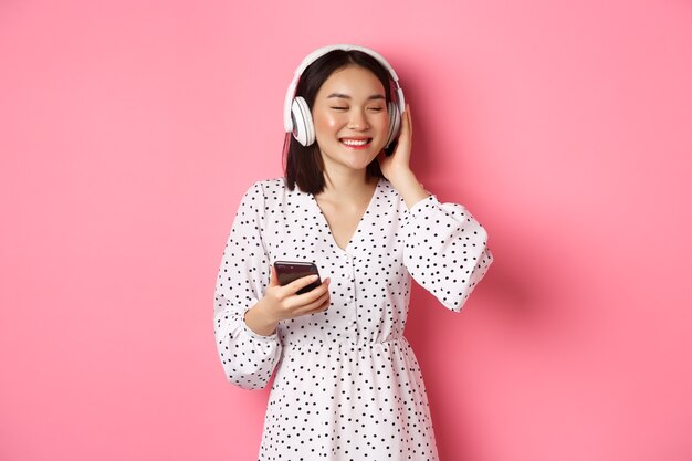 Romantica ragazza asiatica che ascolta musica in cuffia, sorride con gli occhi chiusi, tiene in mano il cellulare, in piedi su sfondo rosa