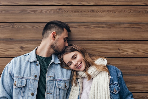 Romantica ragazza bionda sdraiata sulla spalla del ragazzo con gli occhi chiusi. Ritratto dell'interno dell'uomo castana europeo che bacia i capelli della ragazza sulla parete di legno.