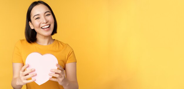 Romanticismo e san valentino felice bella donna asiatica che tiene grande carta del cuore e sorridente in piedi o