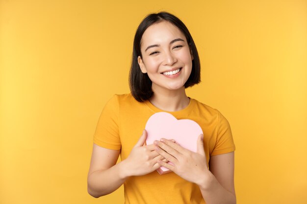 Romanticismo e San Valentino Felice bella donna asiatica che tiene una grande carta del cuore e sorridente in piedi su sfondo giallo