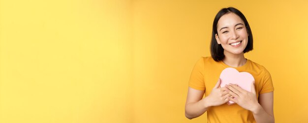 Romanticismo e San Valentino Felice bella donna asiatica che tiene una grande carta del cuore e sorridente in piedi su sfondo giallo