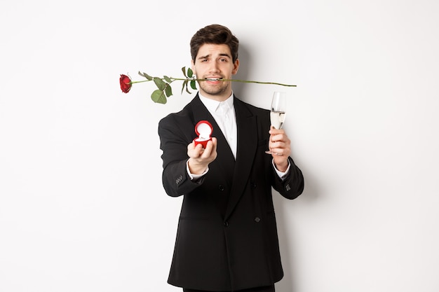 Romantico giovane in abito che fa una proposta, tenendo la rosa tra i denti e un bicchiere di champagne, mostrando l'anello di fidanzamento, chiedendo di sposarlo, in piedi su sfondo bianco.