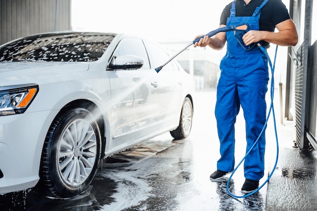 Rondella professionale in uniforme blu che lava auto di lusso con pistola ad acqua su un autolavaggio all'aperto