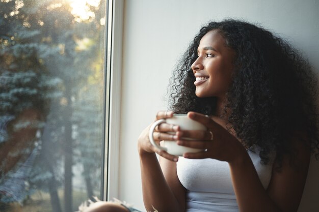 Routine mattutina. Ritratto di felice affascinante giovane donna di razza mista con capelli ondulati godendo di vista estiva attraverso la finestra, bevendo un buon caffè, seduto sul davanzale della finestra e sorridente. Bellissimo sognatore ad occhi aperti