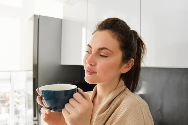 Routine quotidiana e stile di vita giovane bella donna in accappatoio in piedi in cucina con una tazza di caffè