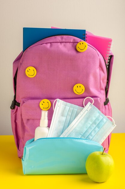 Sacchetto di scuola del bambino di vista frontale con spray per quaderni e maschere sulla scrivania gialla