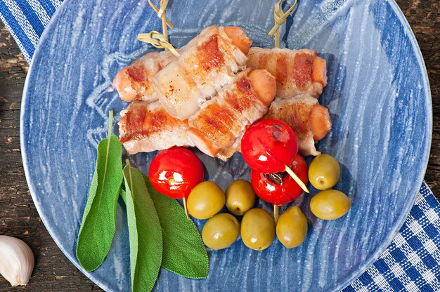 Salsicce alla griglia avvolte in strisce di pancetta con pomodori e foglie di salvia