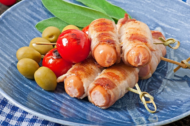 Salsicce alla griglia avvolte in strisce di pancetta con pomodori e foglie di salvia