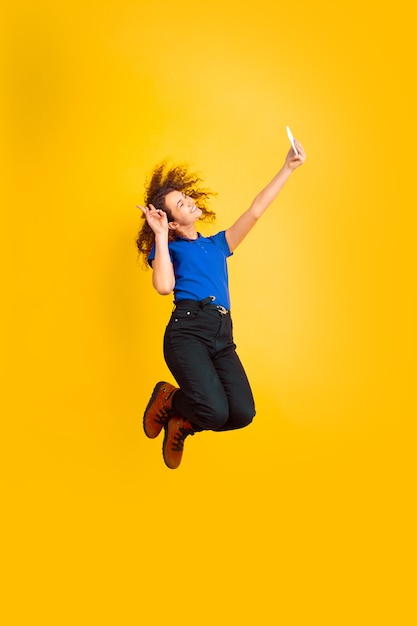 Saltando in alto, prendendo selfie. Ritratto della ragazza dell'adolescente caucasico sulla parete gialla. Bello modello riccio femminile. Concetto di emozioni umane, espressione facciale, vendite, annuncio, educazione. Copyspace.