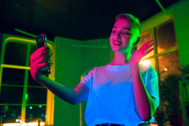 Saluto. Ritratto cinematografico di donna alla moda in interni illuminati al neon. Tonica come effetti cinematografici, colori luminosi al neon. Modello caucasico utilizza lo smartphone in luci colorate al chiuso. Cultura giovanile.