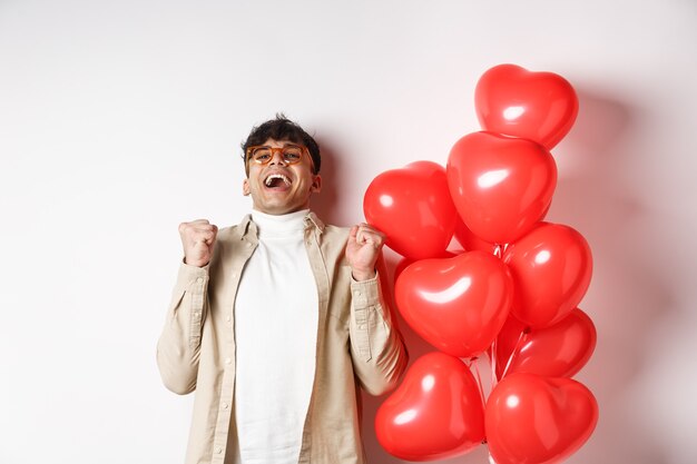 San Valentino. Felice uomo moderno che celebra, urla di gioia e felicità, ha appuntamento con l'amante, è innamorato, in piedi vicino a palloncini a cuore su sfondo bianco