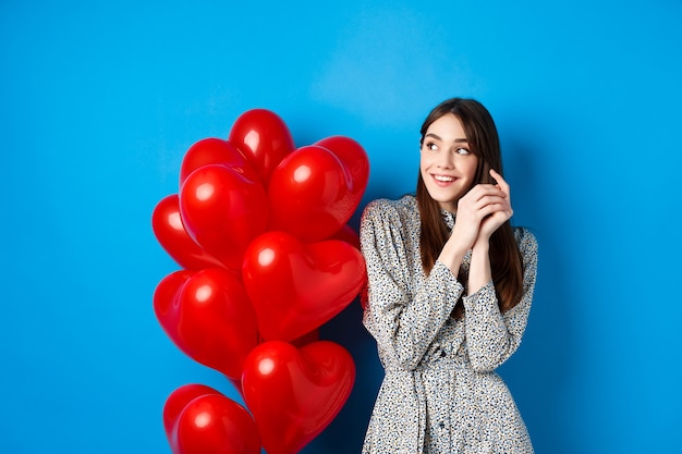 San Valentino. Romantica giovane donna in abito, guardando a sinistra sognante e sorridente, in piedi vicino a palloncini di cuori rossi, sfondo blu