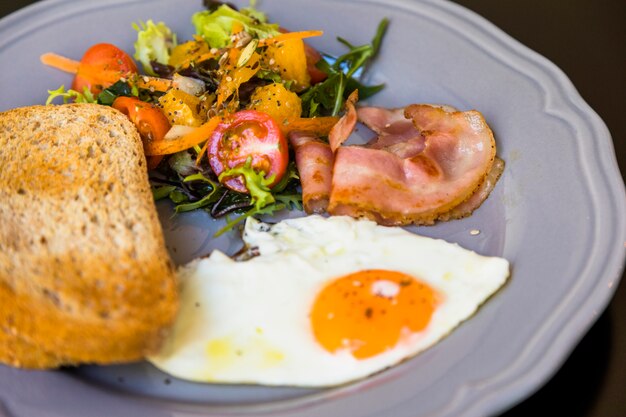 Sana colazione fresca con insalata; Bacon; mezzo uovo fritto e pane tostato sul piatto grigio