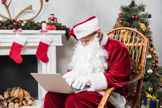 Santa seduto sulla sedia a dondolo e digitando sul computer portatile