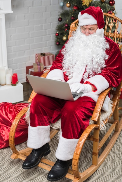 Santa seduto sulla sedia a dondolo e lavorando sul portatile