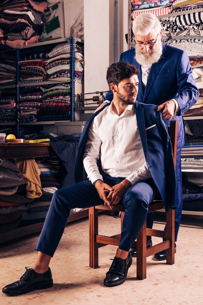 Sarto maschio che prova il cappotto blu sul suo cliente che si siede sopra la sedia di legno nel negozio