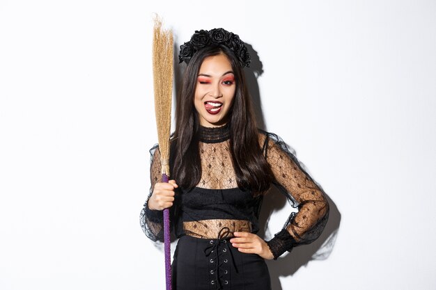 Sassy bella donna asiatica in costume da strega che mostra la lingua, tenendo la scopa e in posa su sfondo bianco.