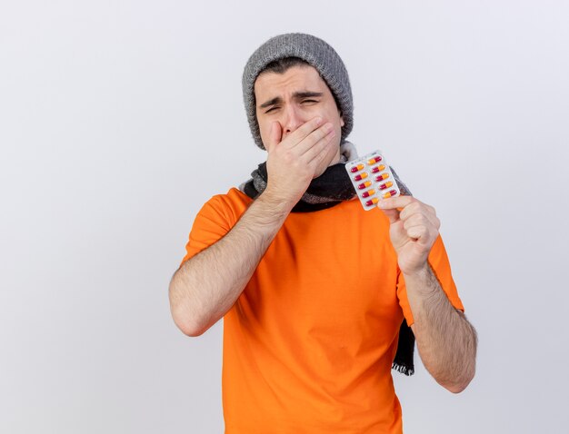 Sbadigliare giovane uomo malato che indossa il cappello invernale con sciarpa tenendo le pillole coperto la bocca con la mano isolata su bianco