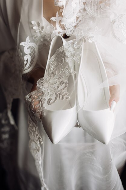 Scarpe da cerimonia bianche da sposa nelle mani della sposa vestite in indumenti da notte di seta con pizzo