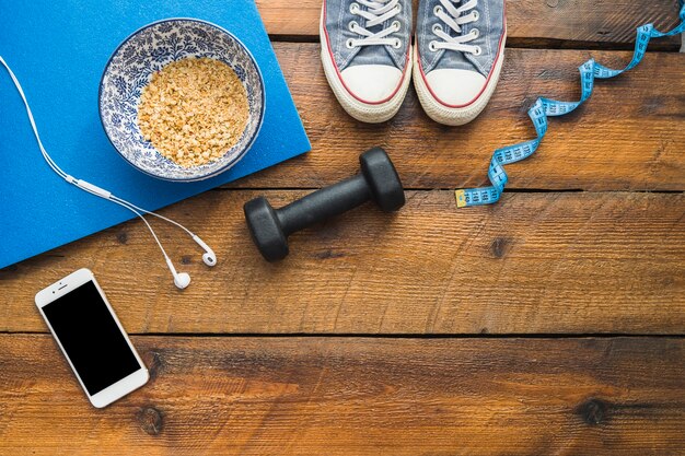 Scarpe; nastro di misurazione; manubrio; auricolare; cellulare e ciotola di avena sul tavolo strutturato in legno