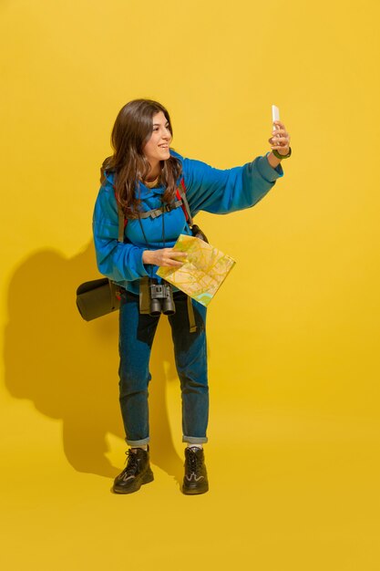 Scattare selfie o vlog. Ritratto di una giovane ragazza turistica caucasica allegra con borsa e binocolo isolato su sfondo giallo studio. Prepararsi per il viaggio. Resort, emozioni umane, vacanze.