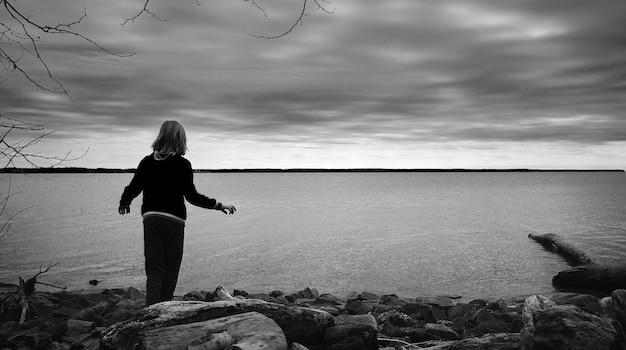 Scatto in scala di grigi di un bambino in piedi sulle rocce in riva al mare e godersi il bellissimo orizzonte calmo