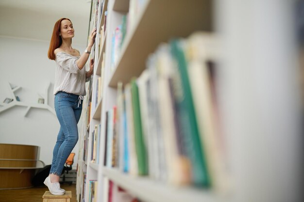 Scegli libro. Giovane donna sorridente dai capelli lunghi snella in camicetta e jeans in cerca di libro in piedi toccando le mani allo scaffale in biblioteca