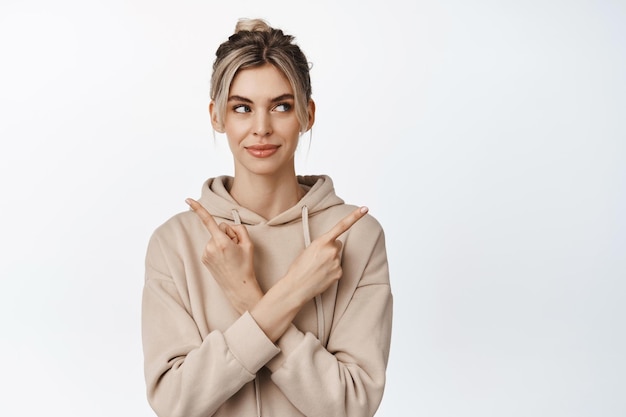 Scelta Donna bionda sorridente che punta le dita lateralmente sbirciando da parte con un sorriso premuroso in piedi su sfondo bianco