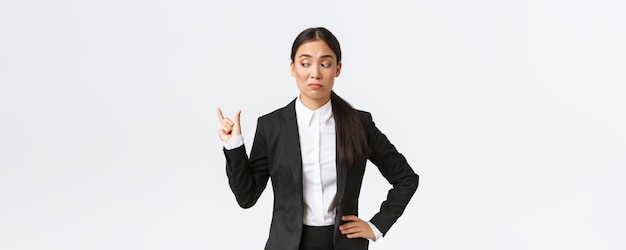 Scettica e goffa giovane donna d'affari asiatica commessa in abito nero che modella qualcosa di piccolo e sembra delusa dalle dimensioni facendo una smorfia disinteressata su una piccola cosa sullo sfondo bianco