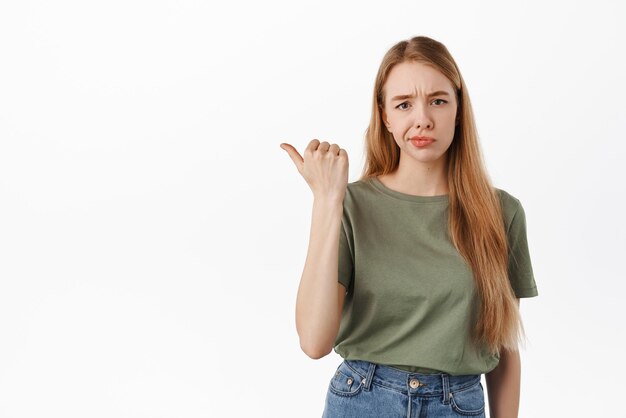 Scettico e dubbioso giovane donna che punta il dito a sinistra ghigno e cipiglio con insoddisfatto volto esitante deluso da qualcosa in piedi su sfondo bianco