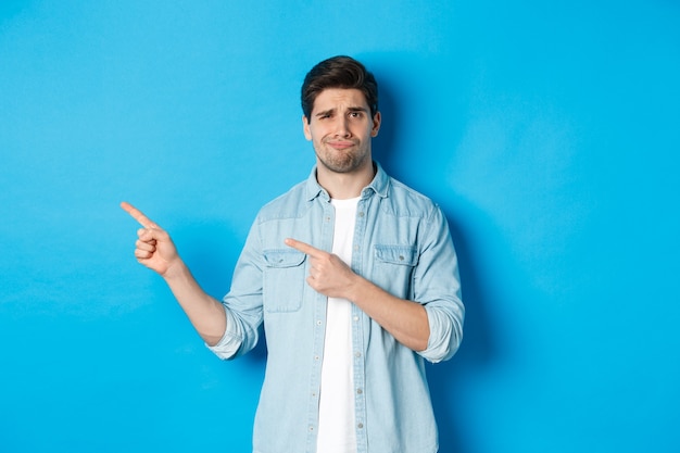 Scettico uomo adulto che punta le dita a sinistra, con aria dubbiosa e incerta, in piedi su sfondo blu