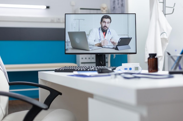 Schermo del computer con medico remoto che ha una conferenza videochiamata online in piedi sul tavolo nell'ufficio dell'ospedale vuoto. Telelavoro in videoconferenza durante la visita medica. Concetto di medicina