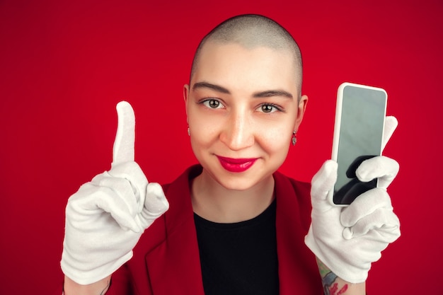 Schermo del telefono vuoto. Ritratto di giovane donna calva caucasica isolata sulla parete rossa dello studio.