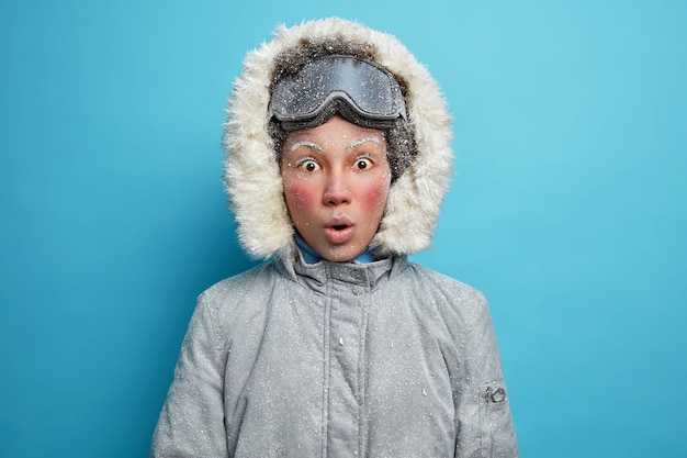 Sciatore femminile scioccato con la faccia rossa congelata guarda vestito con una giacca grigia con cappuccio e occhiali da snowboard.