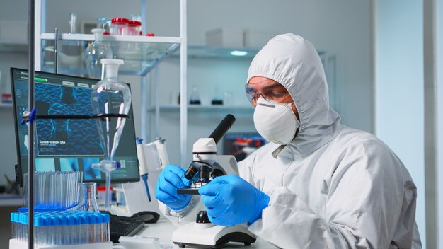Scienziato in tuta dpi posizionando un vetrino sul palco del campione di un microscopio da laboratorio che effettua regolazioni. Chimico in tuta che lavora con vari batteri, campioni di sangue di tessuto per la ricerca sugli antibiotici
