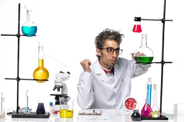 Scienziato maschio di vista frontale in vestito medico che si siede davanti al tavolo con soluzioni su chimica di salute del virus del laboratorio covid scrivania bianca