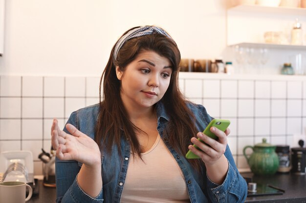 Scioccata emotiva giovane donna sovrappeso che indossa giacca di jeans xxl leggendo un messaggio in cucina a casa con sguardo perplesso durante la navigazione in internet utilizzando il telefono cellulare in cucina durante la colazione