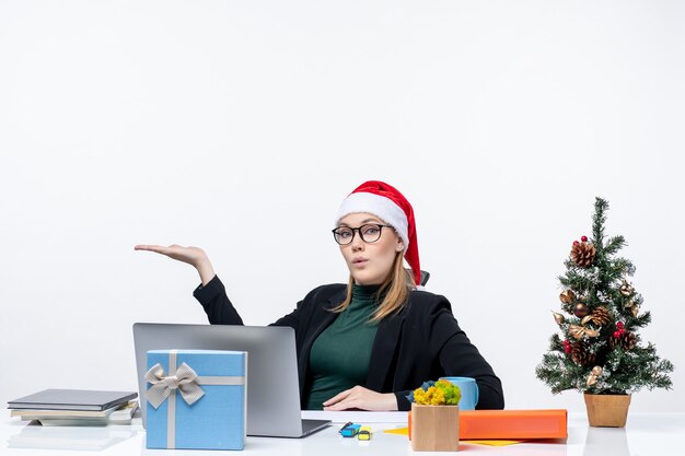 Scioccato giovane donna con cappello di Babbo Natale e occhiali seduti a un tavolo con un albero di Natale e un regalo su di esso su sfondo bianco