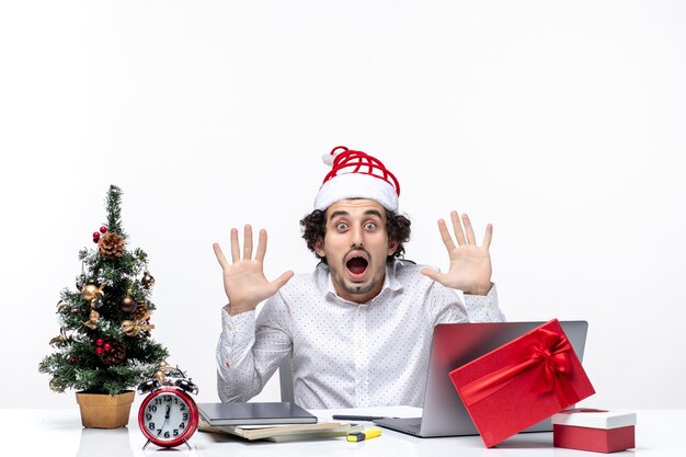 Scioccato giovane imprenditore con buffo cappello di Babbo Natale guardando qualcuno in ufficio su sfondo bianco