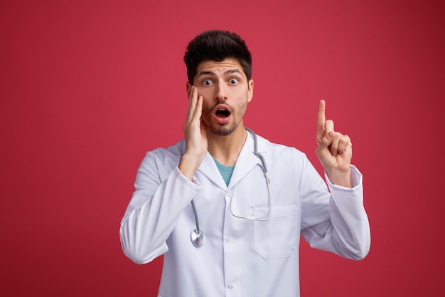 Scioccato giovane medico maschio che indossa uniforme medica e stetoscopio intorno al collo guardando la fotocamera tenendo la mano sul viso rivolto verso l'alto isolato su sfondo rosso