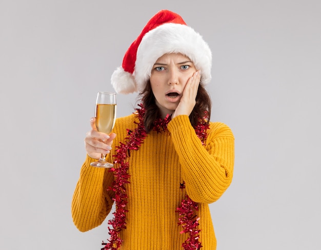 Scioccato giovane ragazza slava con cappello da Babbo Natale e con ghirlanda intorno al collo mette la mano sul viso e tiene un bicchiere di champagne isolato sul muro bianco con spazio di copia