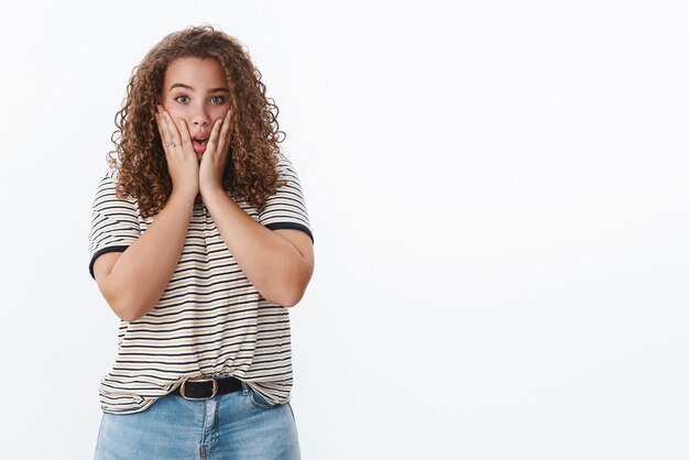 Scioccato preoccupato dai capelli ricci donna grassoccia che reagisce terribili cattive notizie tenere le mani faccia fissando la fotocamera simpatizzando testimone spiacevole evento inquietante in piedi sfondo bianco intenso timido