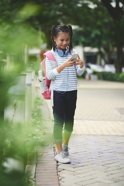 Scolara asiatica adolescente con lo zaino che sta nel parco e che controlla smartphone