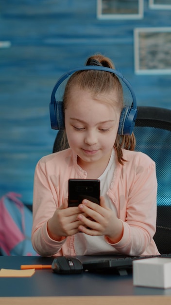 Scolara che utilizza la videochiamata su smartphone per i corsi scolastici a casa. Giovane ragazza che parla con l'insegnante in videoconferenza mentre indossa le cuffie per la lezione online e si siede alla scrivania