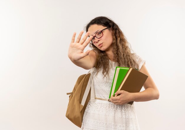 Scolara giovane e carina dispiaciuta con gli occhiali e borsa posteriore che tengono i libri che fanno il gesto di arresto alla macchina fotografica isolata su fondo bianco con lo spazio della copia