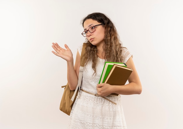 Scolara giovane graziosa confusa con gli occhiali e borsa posteriore che tiene i libri che mostrano la mano vuota che esamina il lato isolato su bianco con lo spazio della copia