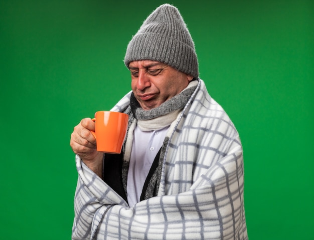 scontento adulto malato uomo caucasico con sciarpa intorno al collo indossando cappello invernale avvolto in plaid guardando la tazza isolata sulla parete verde con spazio di copia