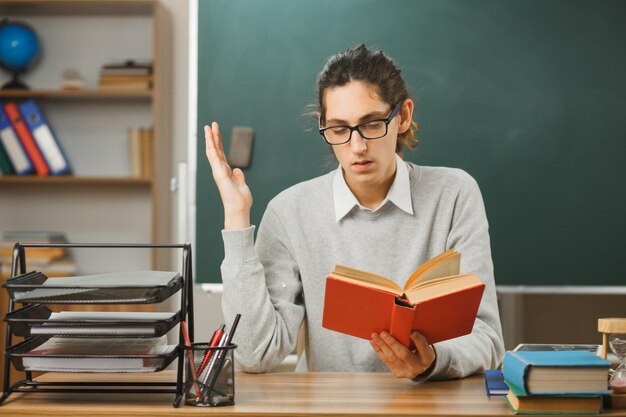 scontento che diffonde mano giovane insegnante maschio che tiene libro seduto alla scrivania con strumenti scolastici in classe